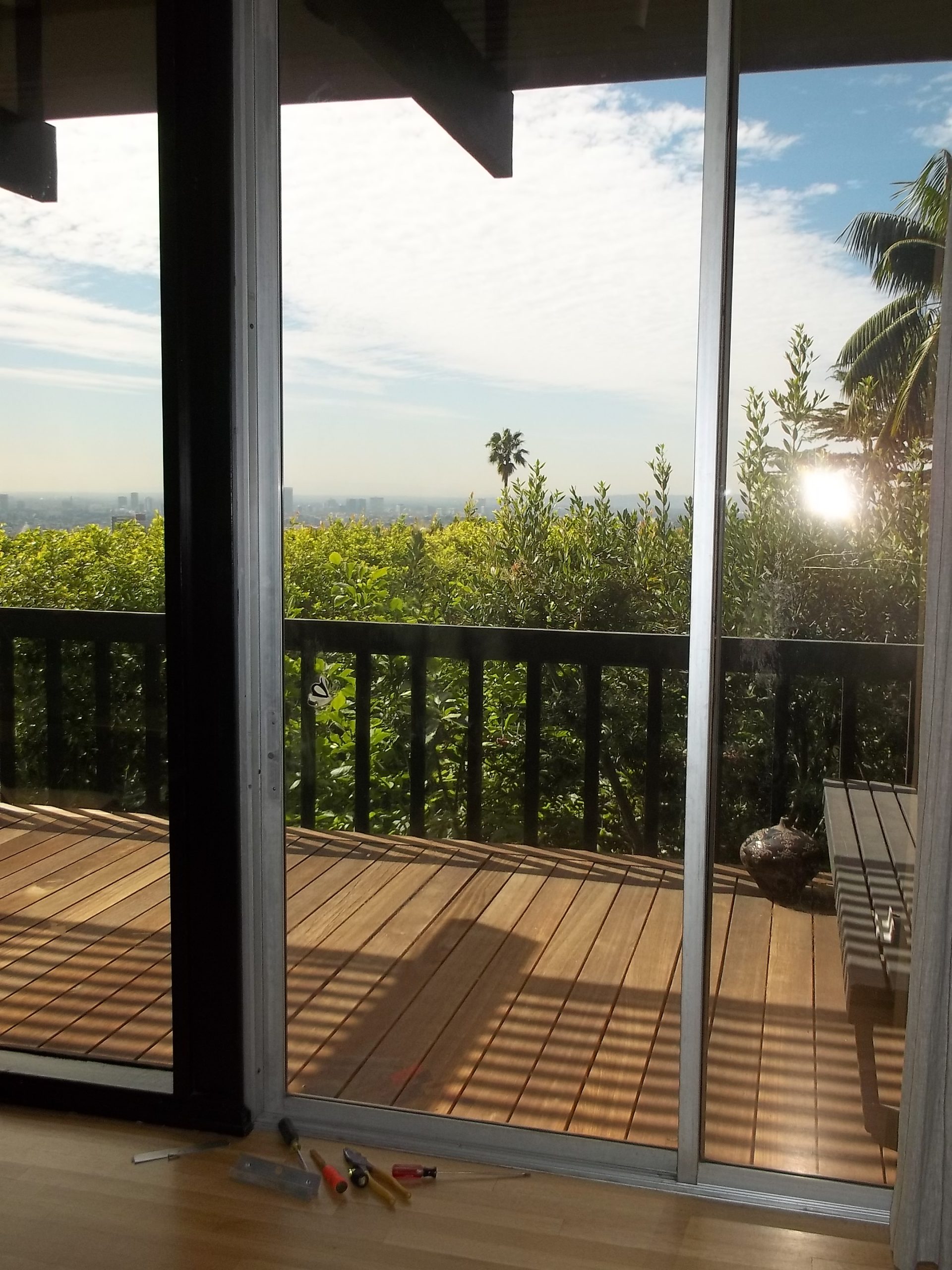 Sliding glass door to wooden balcony or deck overlooking Los Angeles with vegetation growing around it, sun peeking through.